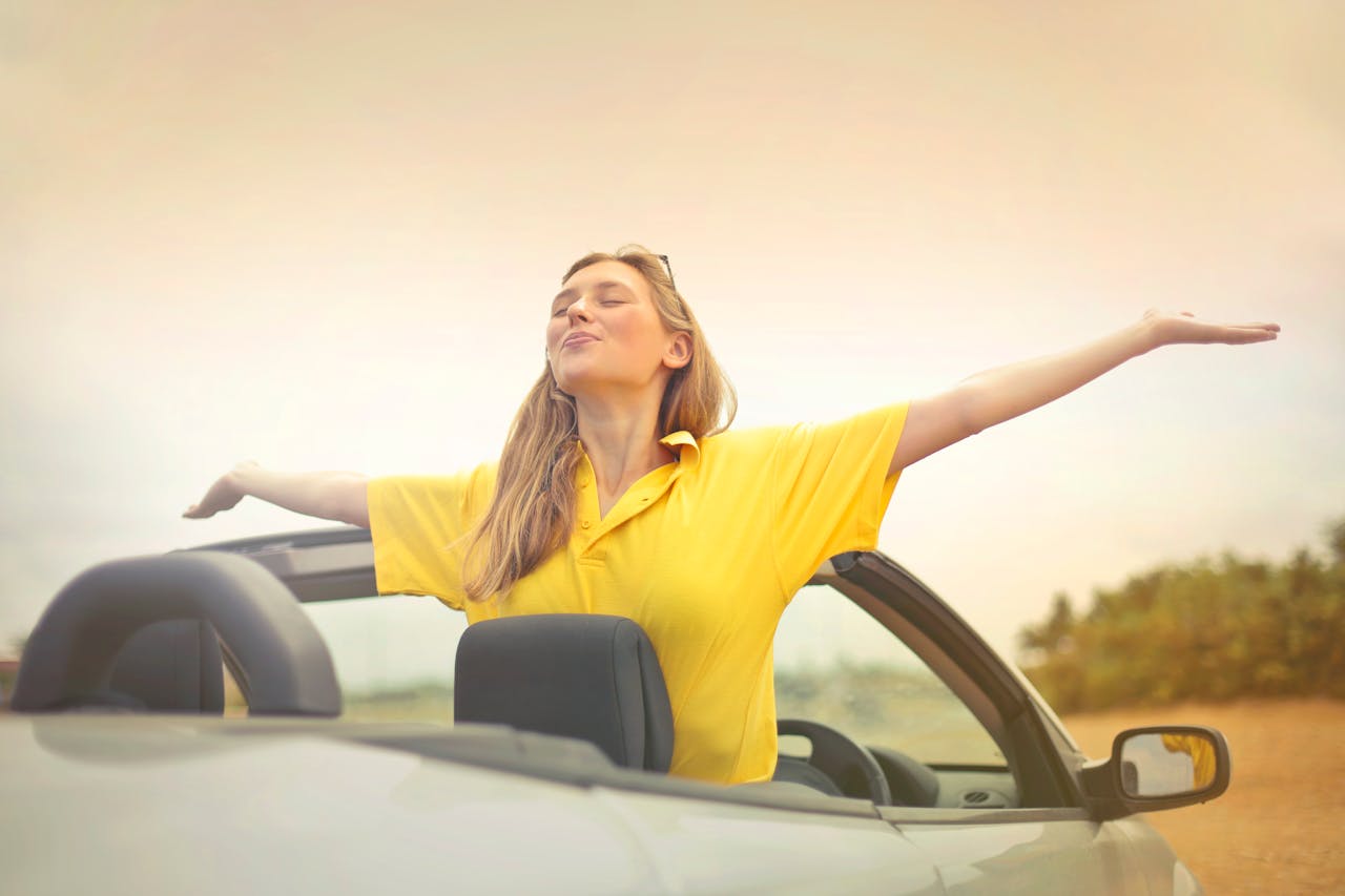 Services A cheerful woman in a yellow shirt enjoys freedom on a sunny drive in a convertible.