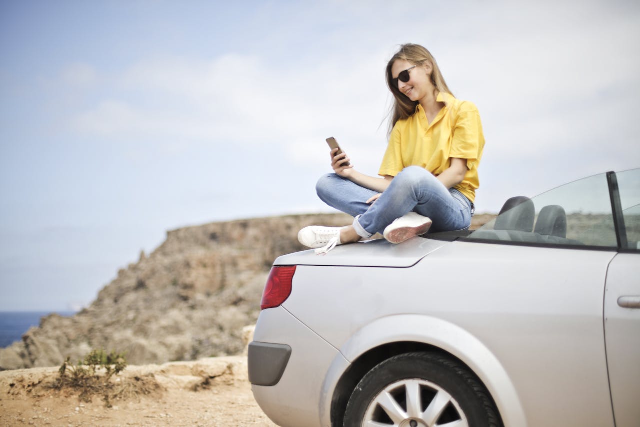 Services Smiling young woman sitting on a car at the beach in Mgarr, Malta using a smartphone.